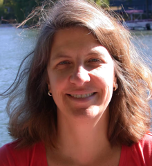  Photo of Dr. Kamlyn Haynes smiling outdoors near a body of water, with shoulder-length brown hair and wearing a red top.