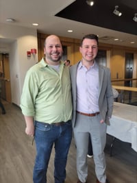 Two men stand arm-in-arm smiling at the camera