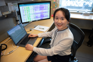 A woman smiling while sitting at a desk with two computer monitors