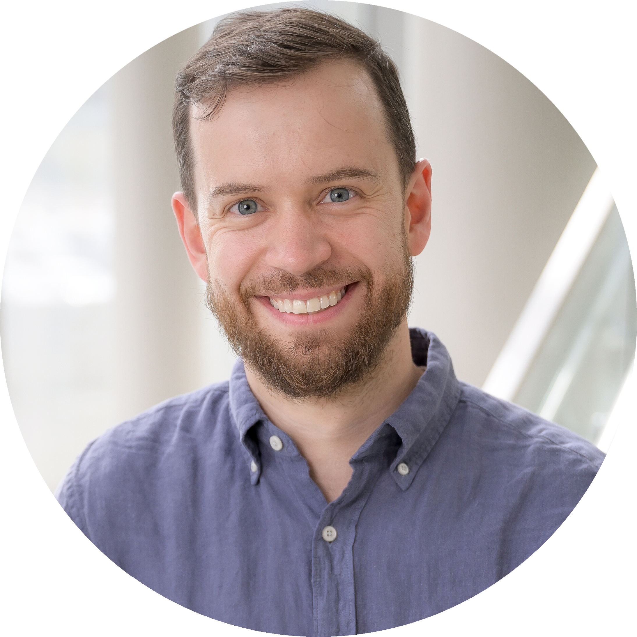 A headshot of Daniel Bondeson, wearing a smile, a beard, and a light blue, button-down shirt. The photo is set indoors with a white modern staircase railing in the background.