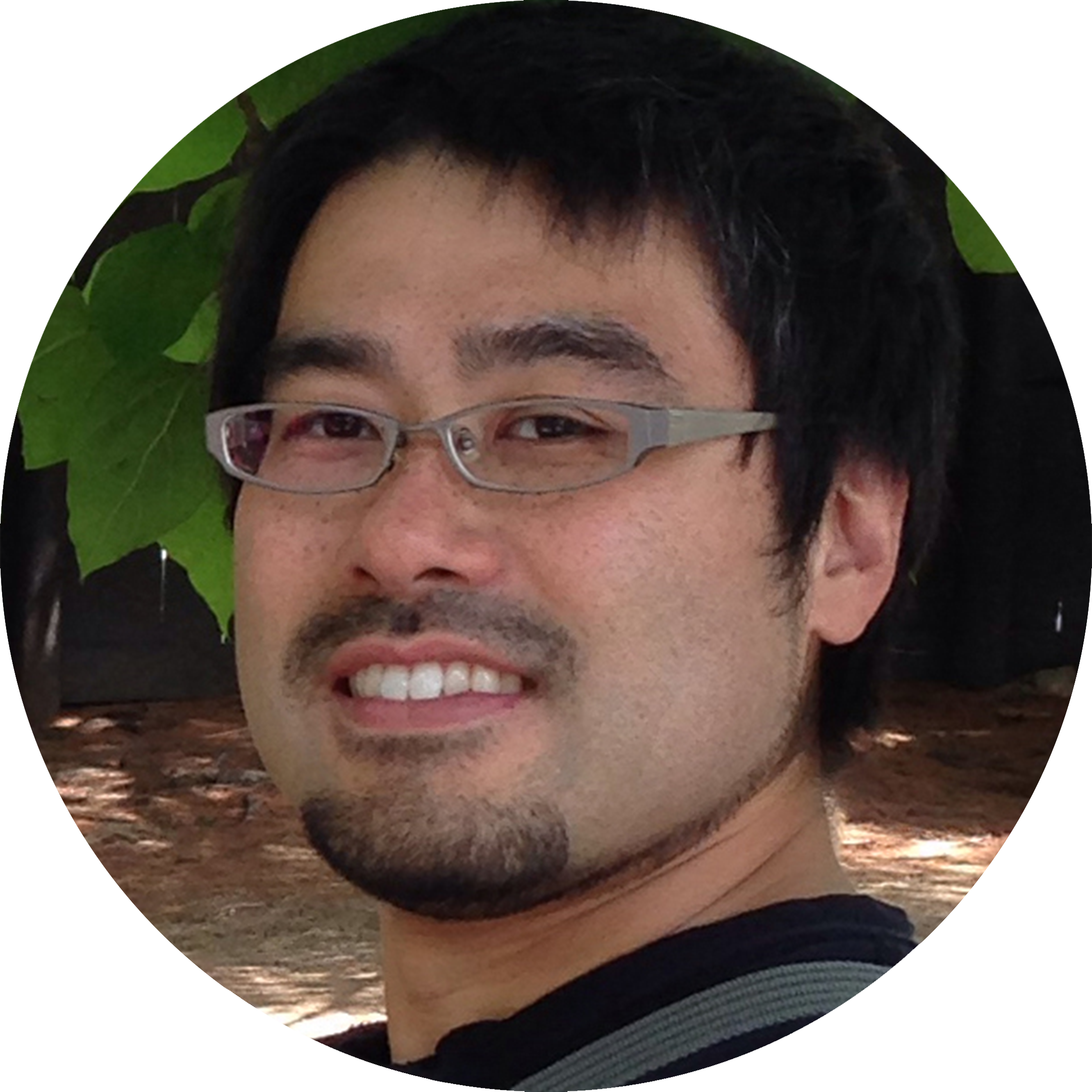 A selfie of Tadatoshi Sato wearing a broad smile in his glasses. He has a short dark hair and a goatee. The photo is set in front of large green leaves as a background.