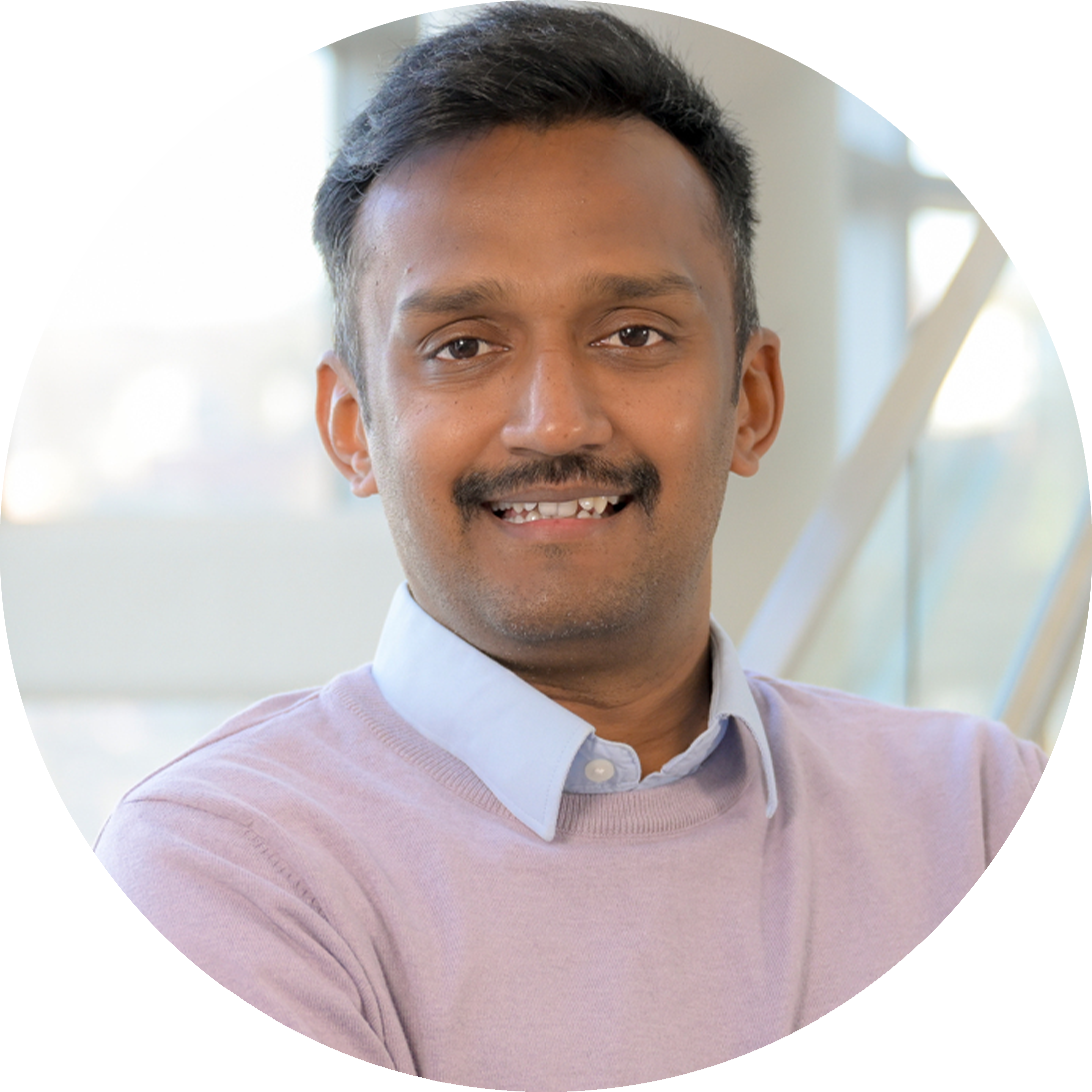 A headshot of Varanasi Siva Karthik, smiling broadly with arms crossed. He is wearing a lavender sweater over a light blue collared shirt, near a staircase with large glass windows in the background.