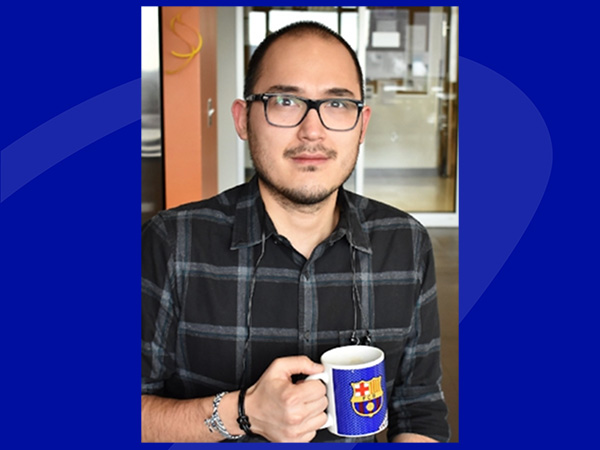 Headshot of Dr Josue Flores Kim, wearing a black boxed shirt with rolled up sleeves, and holding a blue cup with a picture of FC Barcelona logo