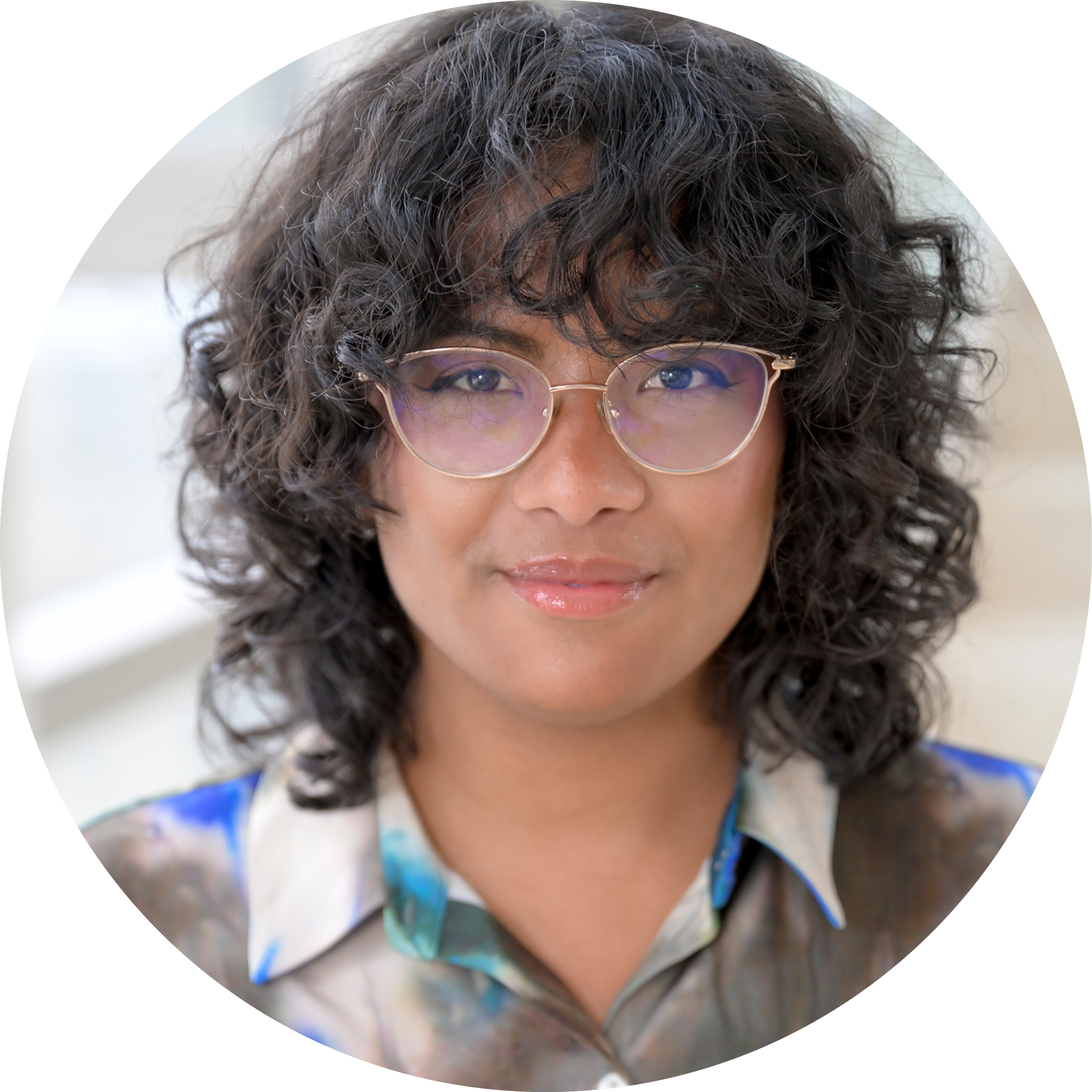 Circular portrait of Natasja Hirabayashi, a mixed-race woman with short-curly brown hair and glasses, wearing a colorful collared shirt