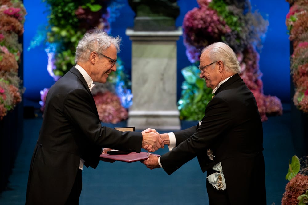 Victor Ambros in a black tuxedo being presented with his Nobel medal by King Carl XVI Gustaf of Sweden.