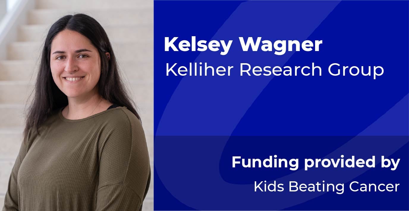 A researcher stands on an indoor staircase beside a blue information panel displaying the text: “Kelsey Wagner, Kelliher Research Group. Funding provided by Kids Beating Cancer.”
