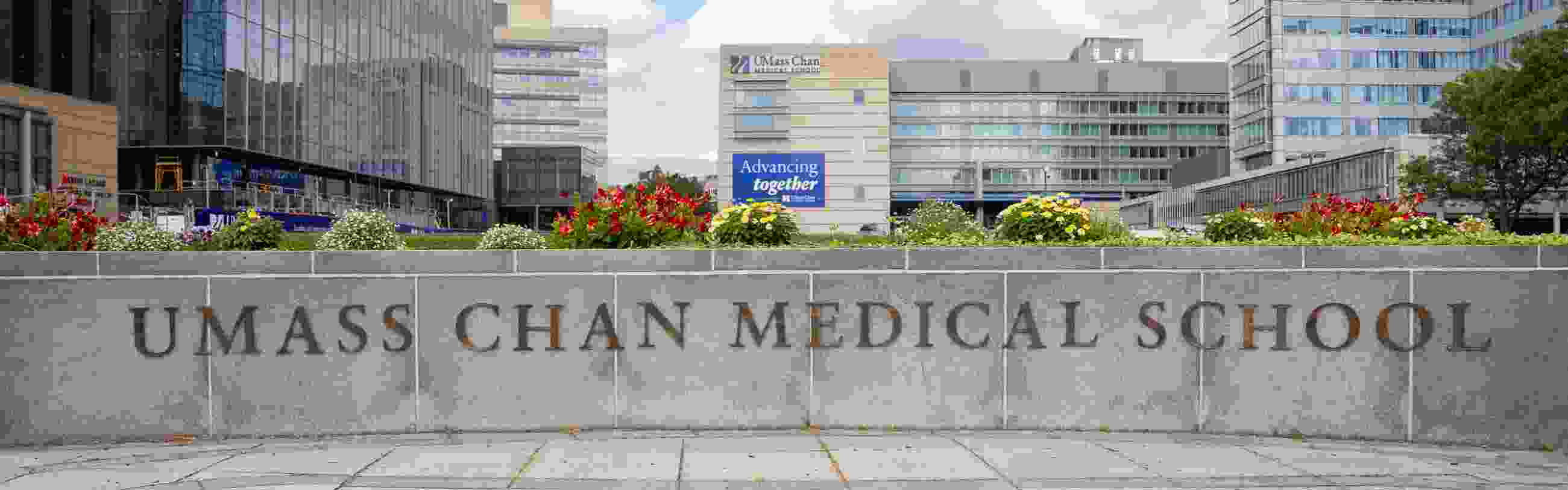 Gold letters on a short wall read "UMass Chan Medical School" with flowers and the campus in the background