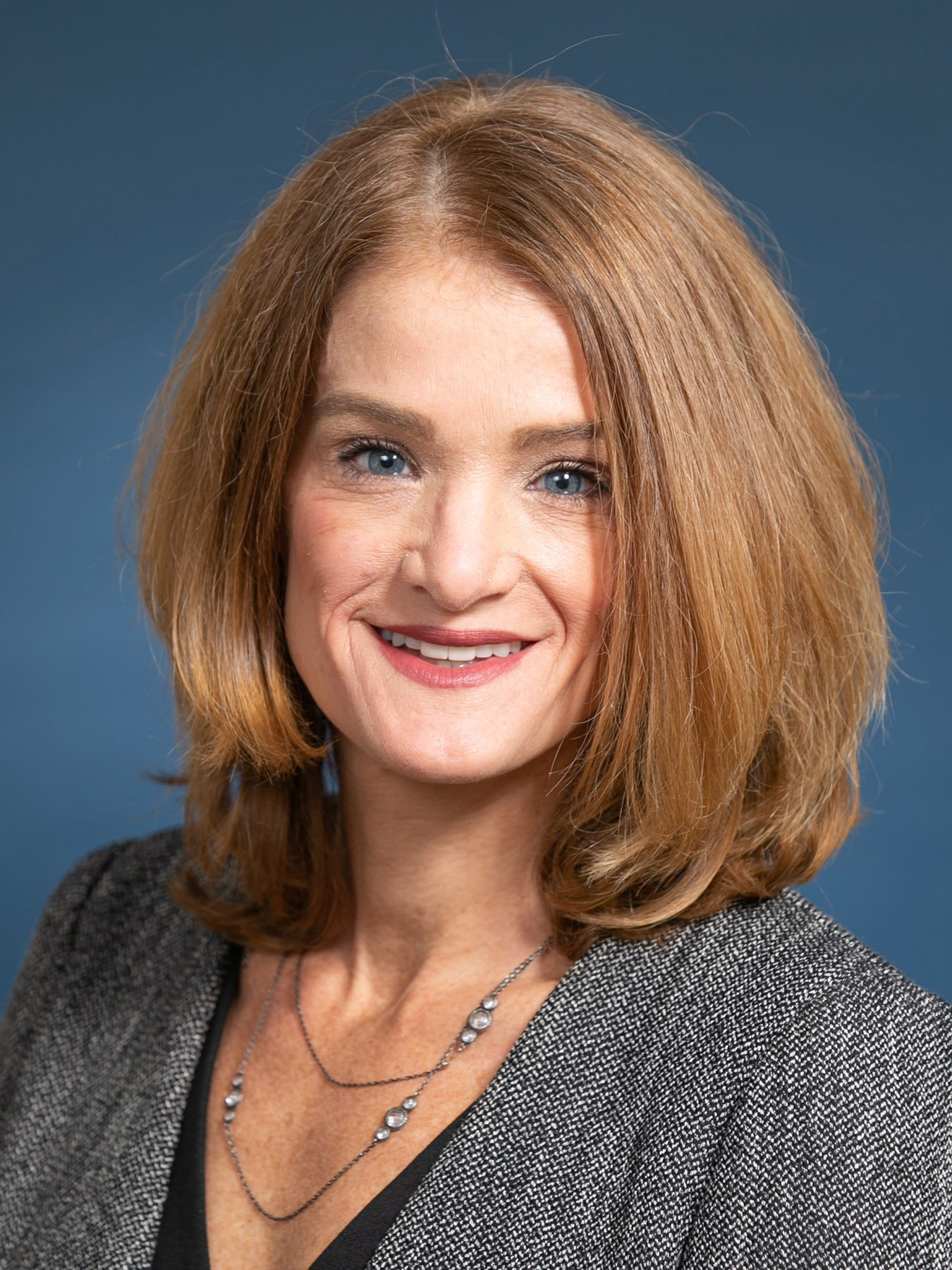 white woman with blue eyes and reddish hair wearing grey jacket smiling