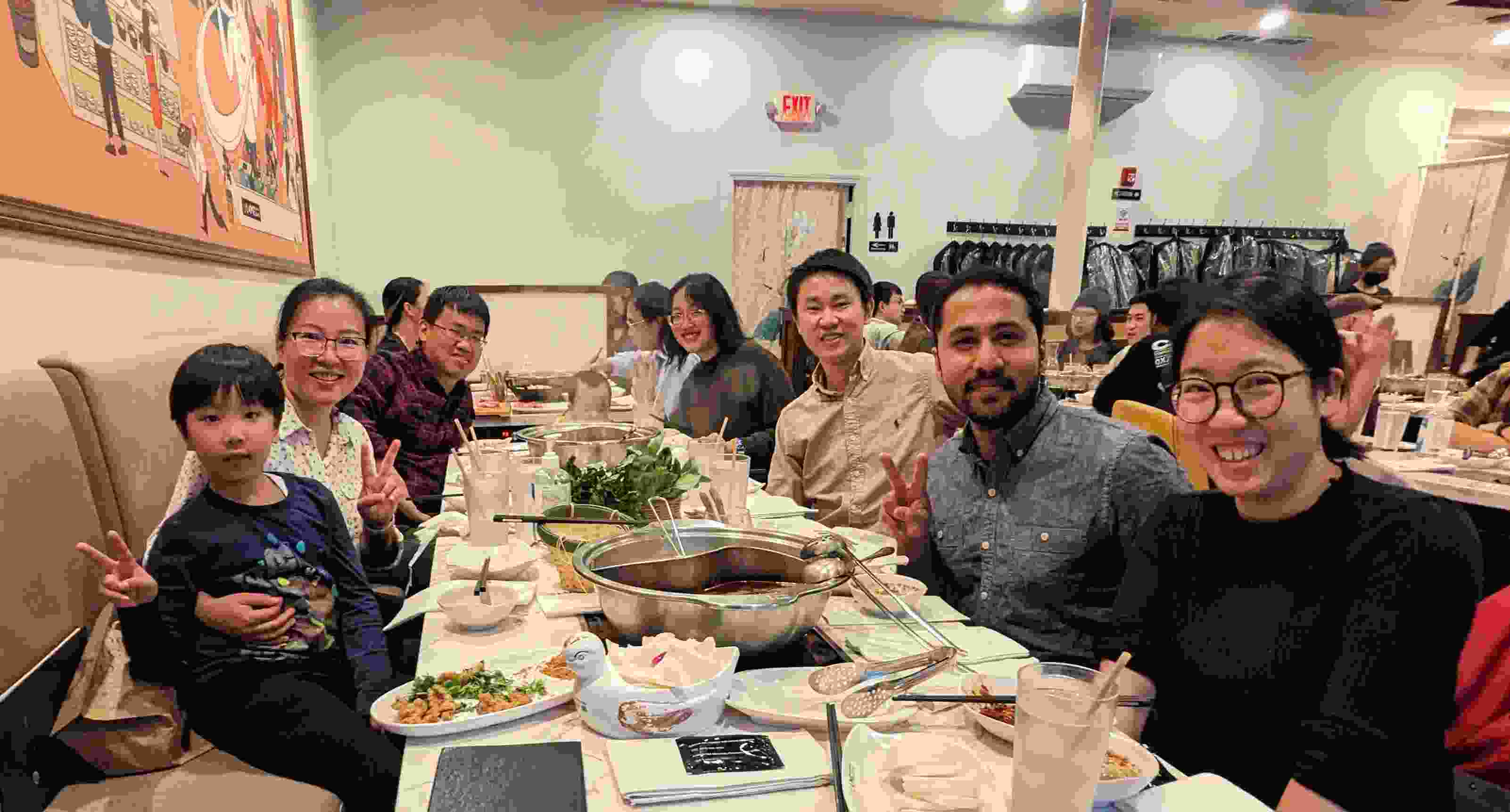 A group of six adults, a kid, and a toddler, sit around a table of two hot pots and food, and smiling together.