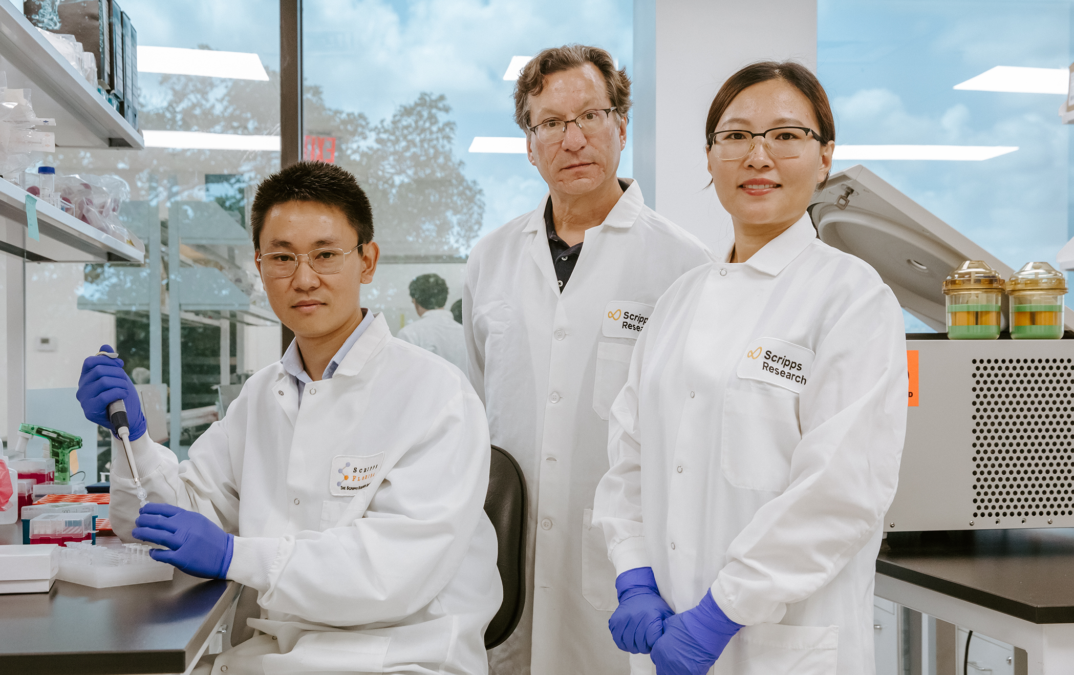 Dr. Zhong sits at a laboratory bench, wearing purple gloves and a white lab coat with a pipette in his hand. Two other scientists stand beside him, also wearing lab coats. 